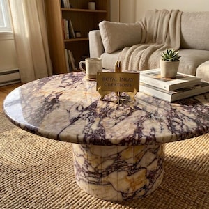 Peut inclure: Une table basse ronde avec une surface polie au motif de marbre crème et violet foncé. La base cylindrique est composée de segments verticaux du même marbre. Un panneau doré porte l'inscription Royal Inlay Creations.