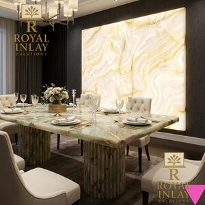 May include: A luxurious dining room featuring a large, rectangular green marble table with cylindrical supports. The table is set with elegant place settings. A large, backlit marble wall panel provides a warm backdrop. The room includes cream-colored chairs and a chandelier.