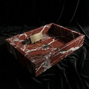 May include: A rectangular red marble sink with white veining. The sink has a polished finish and a circular drain. A small gold-coloured sign with the text "ROYAL INLAY CREATIONS" is attached to the sink.