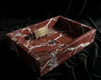 Op de afbeelding: Een rechthoekige rode marmeren wastafel met witte adering. De wastafel heeft een gepolijste afwerking en een ronde afvoer. Een klein goudkleurig bordje met de tekst "ROYAL INLAY CREATIONS" is aan de wastafel bevestigd.