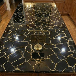 May include: A black countertop with a mosaic pattern of irregular shapes, highlighted by gold-colored veins. A brass sign with the text "ROYAL INLAY CREATIONS" is in the foreground. The countertop is likely made of stone or a similar material.