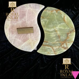 May include: Two decorative onyx pieces, one pink and one green, in a yin-yang design. The pink piece has a light, translucent appearance, while the green piece features brown veining. Each piece has a small, rectangular plaque with the text "ROYAL INLAY CREATIONS".