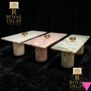 May include: Three rectangular tables with marble tops in cream, pink, and green hues. Each table has a cylindrical base with vertical grooves. The tables are displayed against a black backdrop. The logo "ROYAL INLAY CREATIONS" is visible.