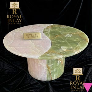 May include: A unique, oval-shaped coffee table with a yin-yang design. The table is crafted from pink and green marble, with a matching cylindrical base. The text "ROYAL INLAY CREATIONS" is displayed on a small gold plaque on the table's surface.