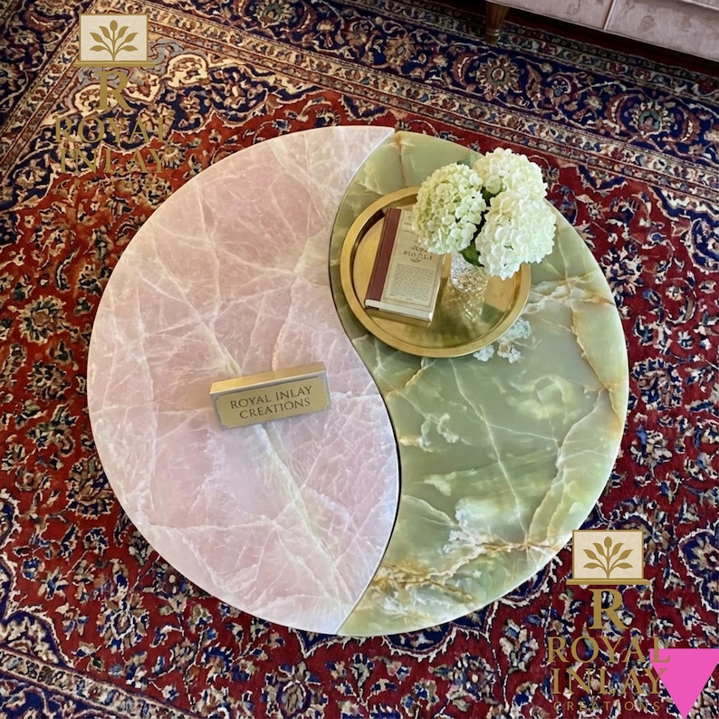 May include: A unique coffee table with a yin-yang design, featuring a pink and green marble top. A gold tray with a book and flowers sits on the green side. The table has the text "Royal Inlay Creations" on a small gold block.