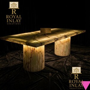 May include: A rectangular, illuminated onyx table with two cylindrical, ribbed supports. The table's surface has a marbled green and brown pattern. The table is lit from within, creating a warm glow. The text "Royal Inlay Creations" is visible.