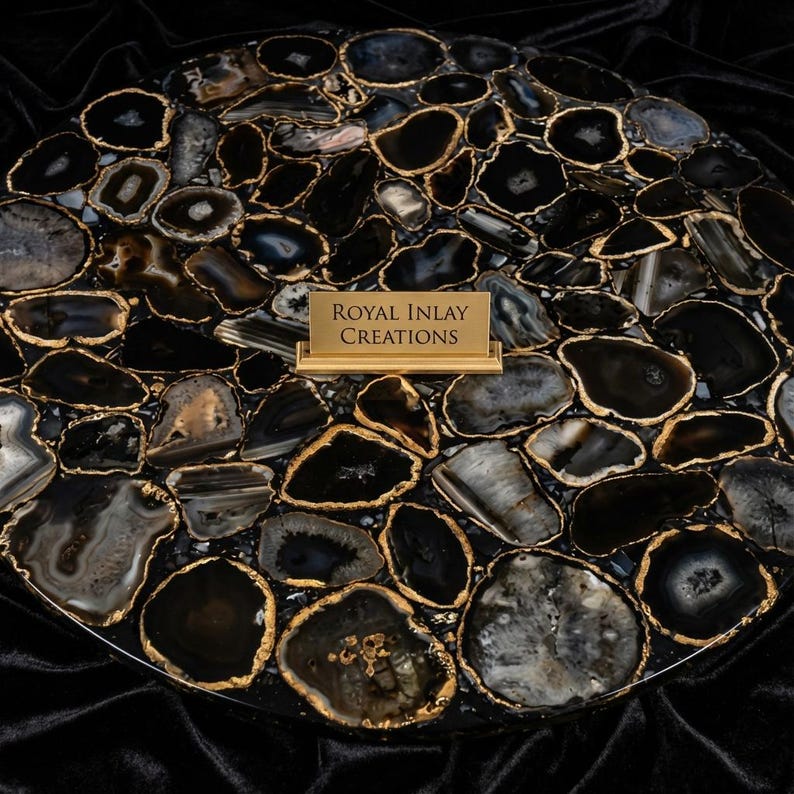 May include: A round, black and gold agate stone table top. The surface is composed of irregularly shaped black and gray agate slices with gold-colored edges. A small gold sign in the center reads "Royal Inlay Creations."
