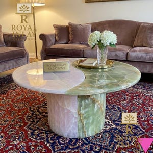 May include: A two-toned coffee table with a pink and green onyx top. The table has a cylindrical base and a yin-yang design. The text "ROYAL INLAY CREATIONS" is visible on the table. A vase of white flowers sits on the table.