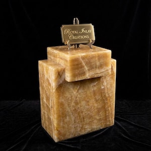 May include: A layered, rectangular sculpture made of golden-brown onyx. A small brass sign with the text "Royal Inlay Creations" sits atop the sculpture. The onyx has natural veining, adding to its unique appearance. The sculpture is displayed against a black background.