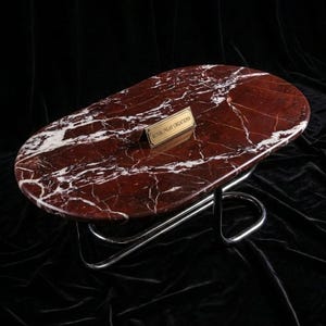 May include: An oval coffee table featuring a polished deep red marble top with prominent white veining. The table is supported by a continuous, curved chrome metal frame. A small rectangular brass plaque on the surface reads ROYAL INLAY CREATIONS.