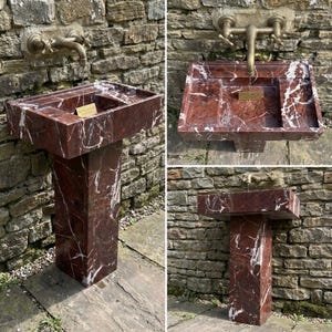 Puede incluir: Un lavabo de m&aacute;rmol rojo con vetas blancas, montado sobre un pedestal a juego. El lavabo tiene una cubeta rectangular con un estante incorporado y se combina con un grifo de lat&oacute;n de estilo vintage. El lavabo est&aacute; colocado contra una pared de piedra.