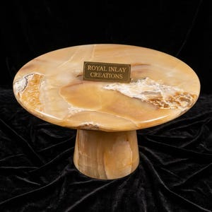 May include: A unique, handcrafted onyx table with a round top and a pedestal base. The table features a warm, earthy color palette with shades of brown, beige, and cream. The text "ROYAL INLAY CREATIONS" is on a small plaque on the table.