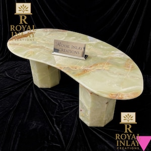Op de afbeelding: Een groene onyx tafel met een uniek, gebogen ontwerp. De tafel heeft een gepolijst oppervlak met bruine adering en wordt ondersteund door twee geometrische, zeshoekige poten. Een zilveren bordje met "Royal Inlay Creations" staat op de tafel.