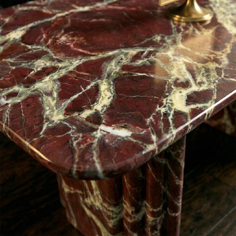May include: A close-up of a rectangular table with a polished, red marble top. The marble features a complex pattern of white and green veining. The table has a matching red marble base.