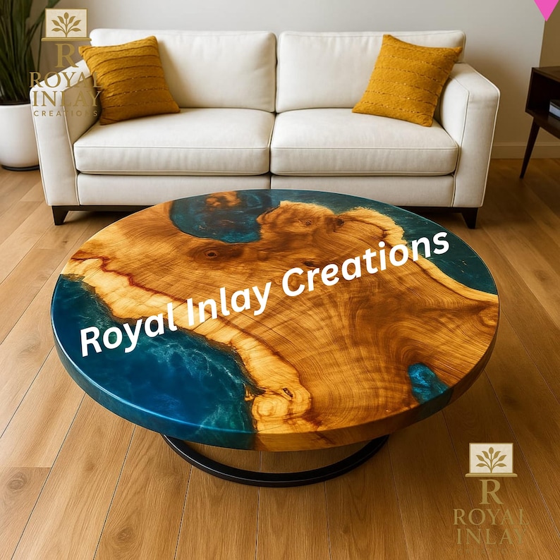May include: Round coffee table with a unique wood grain design and blue resin accents. The table has a dark metal base and the text "Royal Inlay Creations" is visible on the surface. The table is in front of a white sofa with yellow pillows.