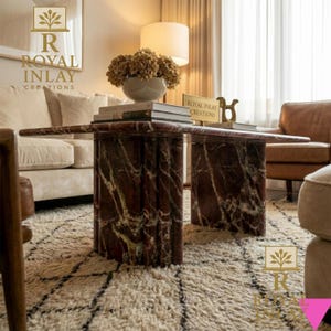 May include: A rectangular coffee table with a red marble top and cylindrical legs. The table is in a living room setting with a cream-colored sofa, a patterned rug, and a lamp.