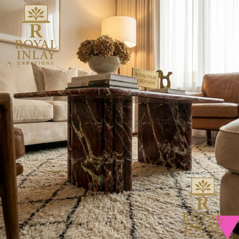 May include: A rectangular coffee table with a red marble top and cylindrical legs. The table is in a living room setting with a cream-colored sofa, a patterned rug, and a lamp.