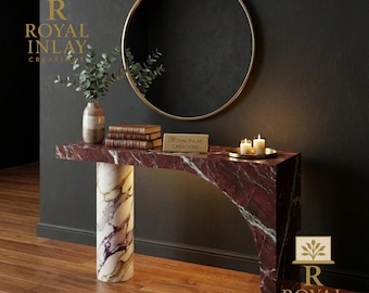 May include: A modern console table with a red marble top and a white and purple marble column base. A gold-framed mirror hangs above the table. The table is decorated with books, a vase with greenery, candles, and a small sign that reads "Royal Inlay Creations".