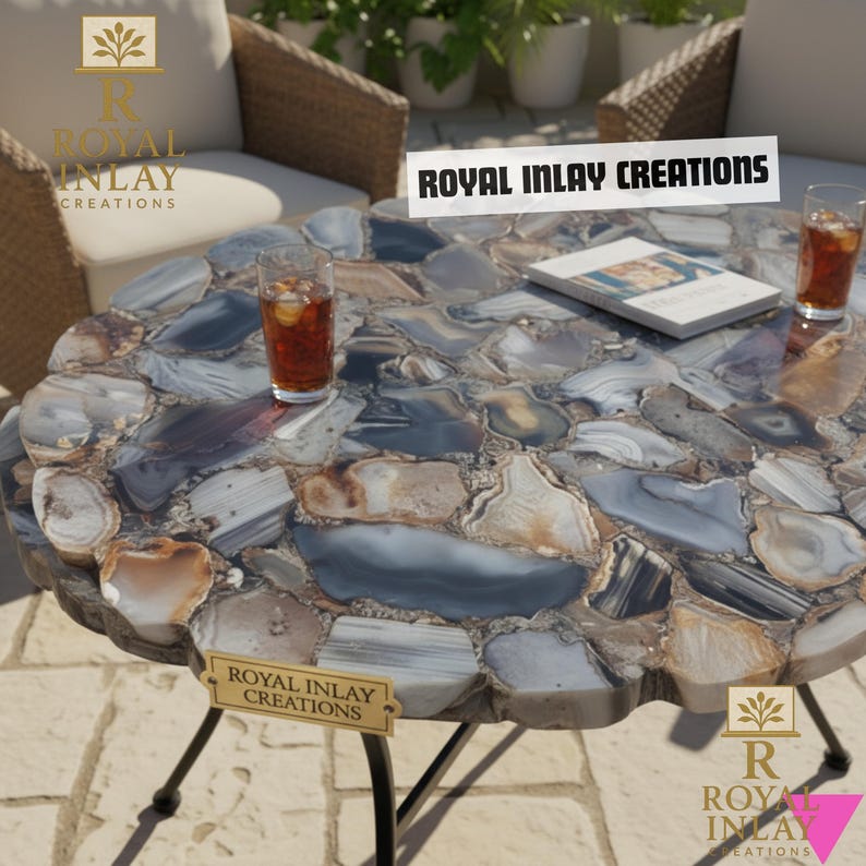 Puede incluir: Una mesa redonda para exteriores con una tapa de mosaico de piedra en tonos grises, azules y marrones. La mesa tiene una base de metal negro y un letrero que dice "ROYAL INLAY CREATIONS". Dos vasos de t&eacute; helado est&aacute;n sobre la mesa.