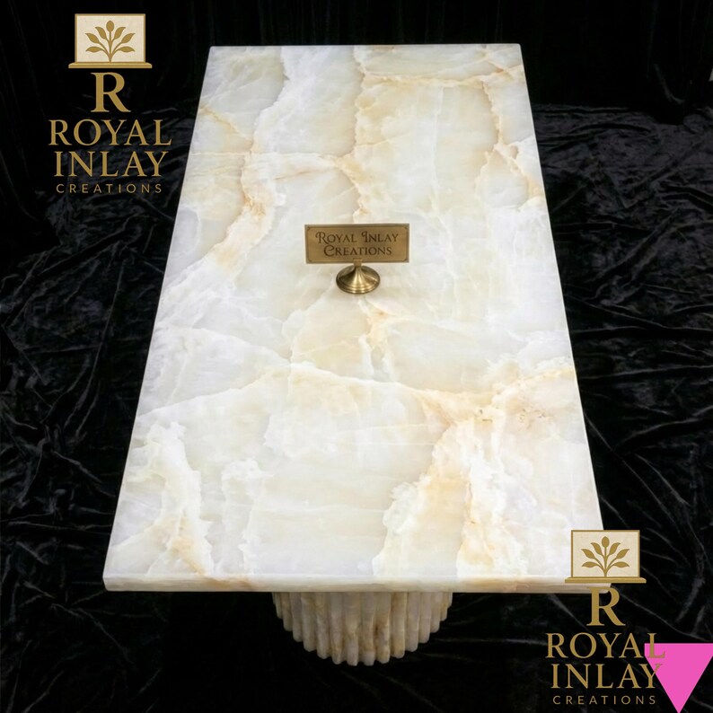 May include: Rectangular white onyx coffee table with a cream and tan veined pattern. The table has a cylindrical base and a small brass plaque that reads "Royal Inlay Creations". The table is set against a black background.