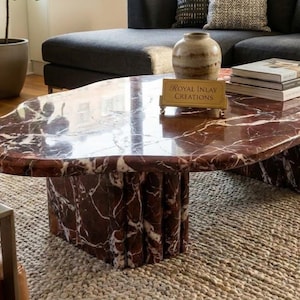 Puede incluir: Una mesa de centro de mármol rojo intenso con vetas blancas, con una superficie de forma orgánica irregular y una base compuesta por columnas de mármol estriadas verticales. Una pequeña placa dorada muestra el texto Royal Inlay Creations.