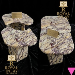 Op de afbeelding: Twee marmeren bijzettafels met een unieke, organische vorm. De tafels hebben een crème- en paars geaderd patroon. Elke tafel heeft een rechthoekige basis en een afgeronde bovenkant. De tekst "Royal Inlay Creations" staat op een kleine gouden plaat op elke tafel.