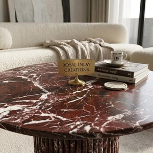 Puede incluir: Una mesa de centro redonda con tapa de m&aacute;rmol rojo y base cil&iacute;ndrica. Un letrero dorado que dice "ROYAL INLAY CREATIONS" est&aacute; sobre la mesa, junto con libros y una taza de t&eacute;.