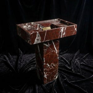 Puede incluir: Un lavabo de m&aacute;rmol rojo con vetas blancas, con una cubeta rectangular y una base de pedestal. El lavabo tiene un peque&ntilde;o estante rectangular y una etiqueta dorada que dice "ROYAL INLAY". El lavabo est&aacute; sobre un fondo oscuro.