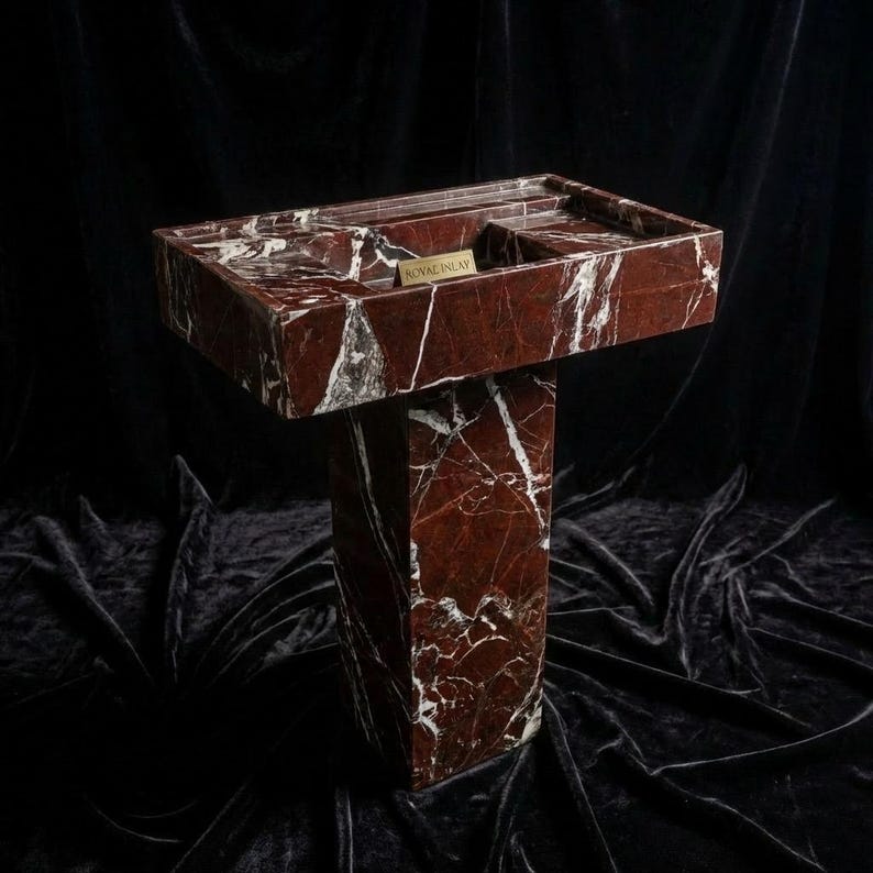 Puede incluir: Un lavabo de m&aacute;rmol rojo con vetas blancas, con una cubeta rectangular y una base de pedestal. El lavabo tiene un peque&ntilde;o estante rectangular y una etiqueta dorada que dice "ROYAL INLAY". El lavabo est&aacute; sobre un fondo oscuro.