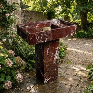 Puede incluir: Un lavabo exterior de m&aacute;rmol rojo con vetas blancas, colocado sobre un pedestal a juego. El lavabo tiene una cubeta rectangular con una peque&ntilde;a abertura rectangular. El lavabo est&aacute; rodeado de exuberante vegetaci&oacute;n y un camino de piedra. Una peque&ntilde;a placa dorada dice "Royal Inlay Creations."