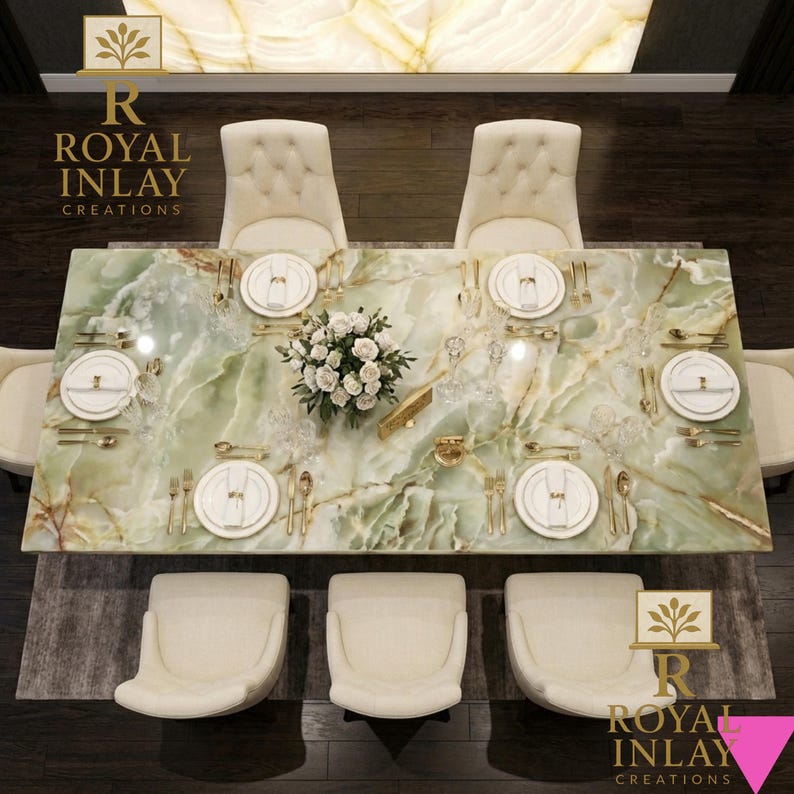 May include: Overhead view of a dining table with a green marble top, set for a meal. The table is surrounded by cream-colored chairs. Gold-colored cutlery and white plates are arranged on the table, along with a floral centerpiece.