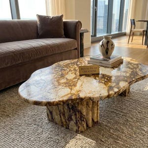 May include: An oval coffee table with a polished brown and white marble top and a fluted marble base. A small rectangular plaque on the surface displays the text ROYAL INLAY CREATIONS. The table features natural veining throughout the stone surface.
