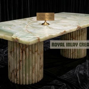 May include: A rectangular onyx table with a green and brown marbled surface. The table rests on two cylindrical bases with vertical ribbed detailing. The text "Royal Inlay Creations" is visible on the table and in the logo.