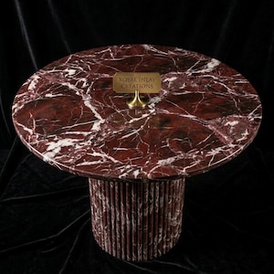 May include: A round marble table with a deep red and white veined top. The base is cylindrical, made of vertical red and white marble pieces. A small brass sign reads "ROYAL INLAY CREATIONS".
