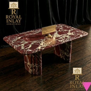 May include: A rectangular marble coffee table with a red and white veined pattern. The table has a matching base and a small gold sign that reads "Royal Inlay Creations". The table is on a dark wood floor.