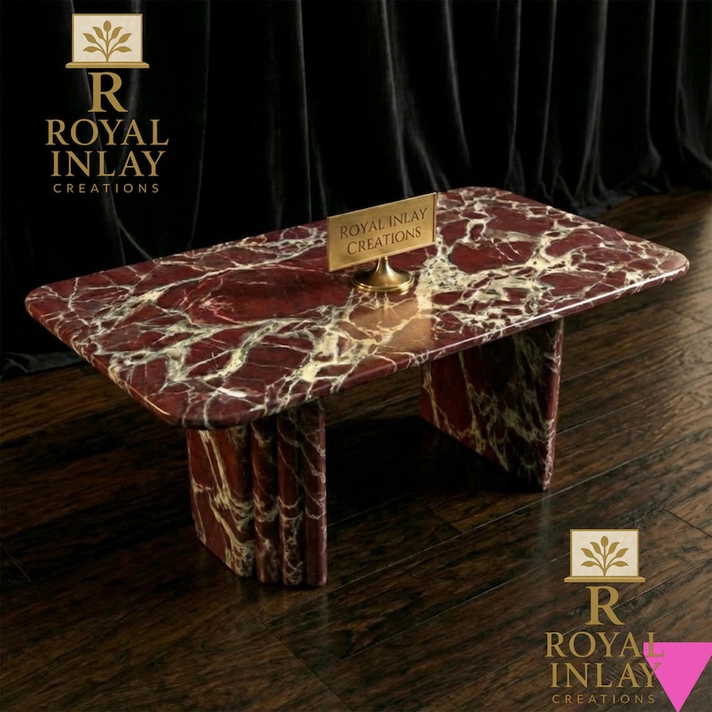May include: A rectangular marble coffee table with a red and white veined pattern. The table has a matching base and a small gold sign that reads "Royal Inlay Creations". The table is on a dark wood floor.