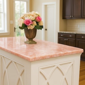 Puede incluir: Isla de cocina con encimera rosa y gabinetes blancos. Un arreglo floral en un jarr&oacute;n decorativo se encuentra sobre la encimera. La cocina cuenta con gabinetes de madera oscura y electrodom&eacute;sticos de acero inoxidable.