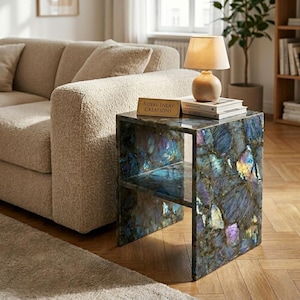 Puede incluir: Una mesa auxiliar cuadrada construida con losas de piedra labradorita iridiscente que presentan inclusiones minerales en azul profundo, morado y dorado. La mesa tiene un diseño de estante abierto y una placa rectangular con el texto Royal Inlay Creations.