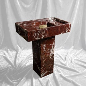 Puede incluir: Un lavabo de pedestal de m&aacute;rmol rojo con vetas blancas. La pila rectangular tiene un borde incorporado y un &aacute;rea rectangular peque&ntilde;a. La base es una columna rectangular s&oacute;lida. El texto "ROYAL INLAY CREATIONS" est&aacute; en una peque&ntilde;a placa dorada.