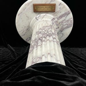 May include: A white marble pedestal with purple veining, featuring a circular top and a fluted cylindrical base. A rectangular wooden plaque with a brass plate is attached to the top, engraved with the text ROYAL INLAY CREATIONS.