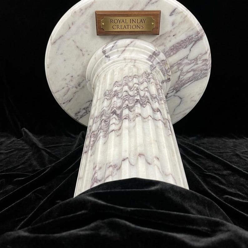 May include: A white marble pedestal with purple veining, featuring a circular top and a fluted cylindrical base. A rectangular wooden plaque with a brass plate is attached to the top, engraved with the text ROYAL INLAY CREATIONS.