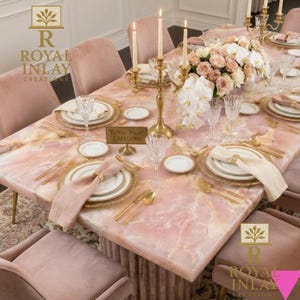 May include: A formal dining table set for a special occasion. The table features a pink marble top, gold-rimmed plates, gold cutlery, and pink napkins. The centerpiece includes a floral arrangement and lit candles. The chairs are upholstered in pink velvet.