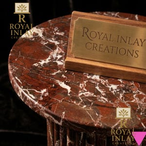 May include: A round, red marble table with white veining. A wooden sign with a gold plaque that reads "Royal Inlay Creations" is on the table. The table has a fluted base and is set against a dark background.