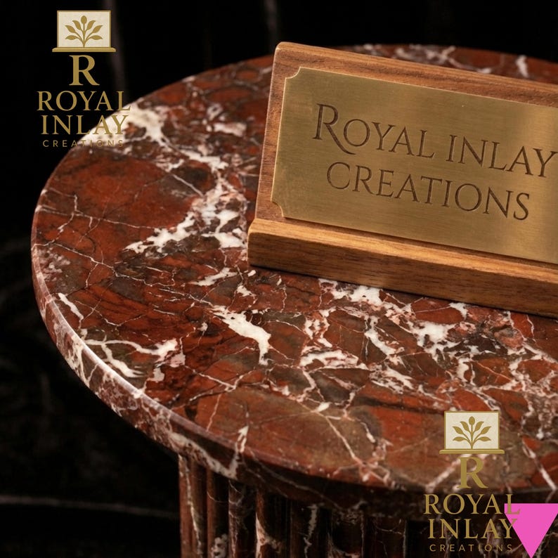 May include: A round, red marble table with white veining. A wooden sign with a gold plaque that reads "Royal Inlay Creations" is on the table. The table has a fluted base and is set against a dark background.