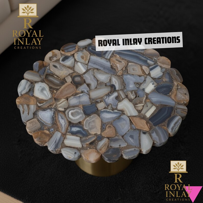Puede incluir: Mesa ovalada con una tapa de mosaico hecha de piedras pulidas de varios tama&ntilde;os, en tonos grises, azules y marrones. La mesa tiene una base dorada y redondeada. El texto "ROYAL INLAY CREATIONS" se muestra en la parte superior.