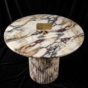 Puede incluir: Mesa ovalada con tapa de mármol y base cilíndrica. La encimera presenta un patrón veteado blanco, morado y dorado. La base está hecha de varillas de piedra verticales y claras. Un pequeño letrero de latón dice "ROYAL INLAY CREATIONS".