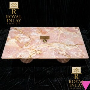 May include: A rectangular pink onyx coffee table with a natural marble pattern. The table has a light pink hue with golden veining and is supported by two cylindrical, textured bases. The table has a small brass sign that reads "Royal Inlay Creations".