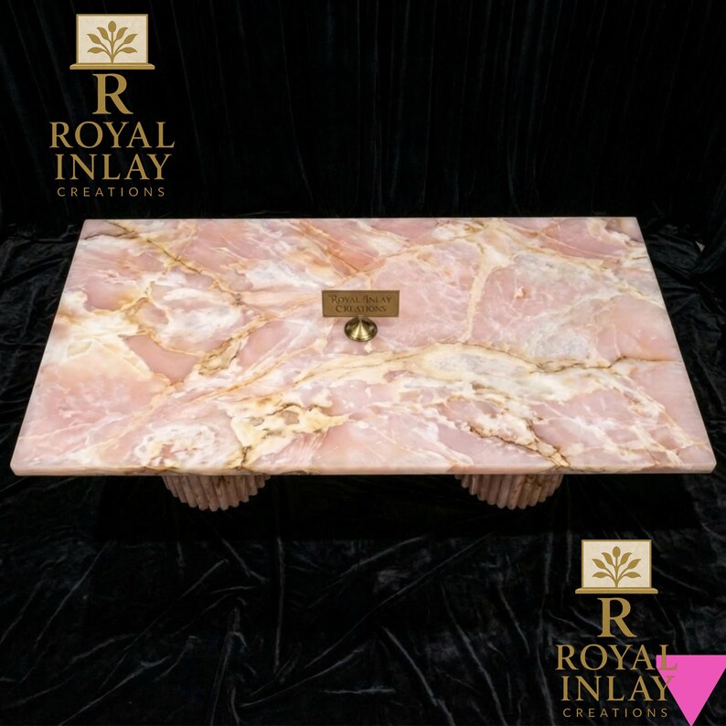 May include: A rectangular pink onyx coffee table with a natural marble pattern. The table has a light pink hue with golden veining and is supported by two cylindrical, textured bases. The table has a small brass sign that reads "Royal Inlay Creations".