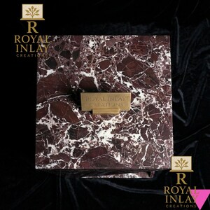 Puede incluir: Una caja cuadrada de m&aacute;rmol con un patr&oacute;n burdeos y blanco. Una placa de metal dorada con el texto "ROYAL INLAY CREATIONS" est&aacute; fijada en la parte delantera. La caja est&aacute; sobre un fondo oscuro, con el logotipo "Royal Inlay Creations" en las esquinas.