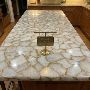 May include: A kitchen countertop with a white and gold stone pattern. The countertop has a polished surface and is made of large, irregular pieces of white stone with gold-coloured veins. A gold-coloured sign that reads "Royal Inlay Creations" is on the countertop.